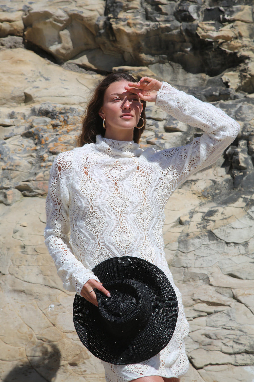 Resort Collection raffia hat held in hand with a white lace blouse by the sea